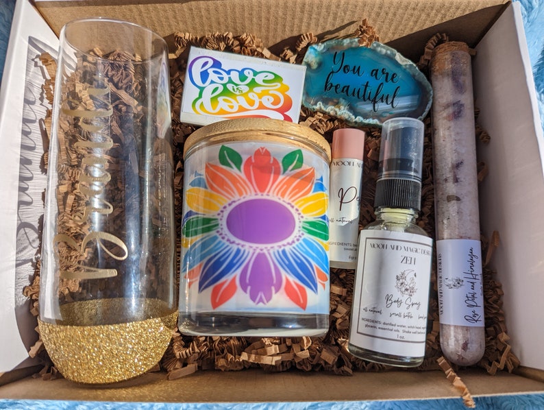May include: A gift box filled with self-care items, including a rainbow candle, a glass with glitter, a bottle of body spray, a tube of bath salts, a sticker that says "Love is Love", and a stone that says "You are beautiful".
