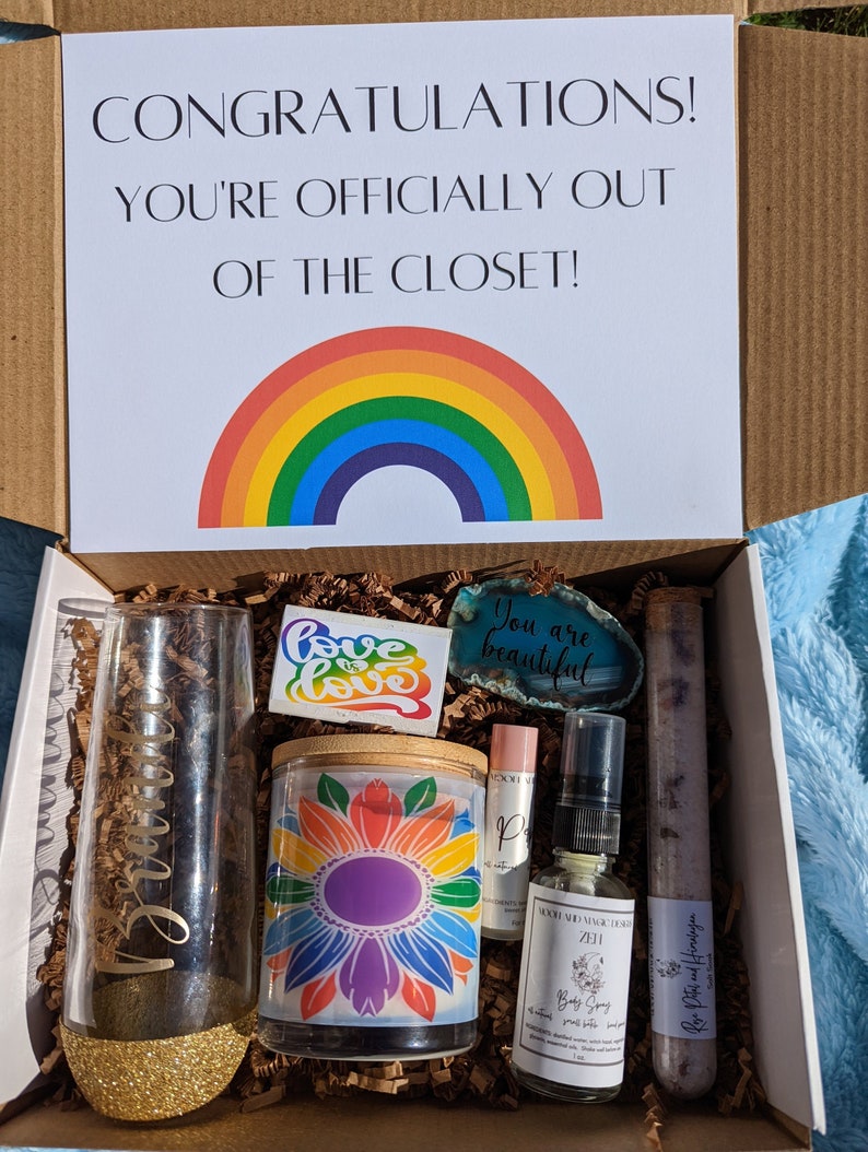 May include: A gift box filled with rainbow-themed items, including a candle, a glass with glitter, a bath bomb, a bottle of body spray, and a stone with the text "You are beautiful". The box lid has a rainbow and the text "Congratulations! You're officially out of the closet!"