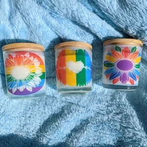 May include: Three glass candle jars with wooden lids. The jars have different designs: a rainbow with a heart, a white sunflower, and a colorful sunflower.