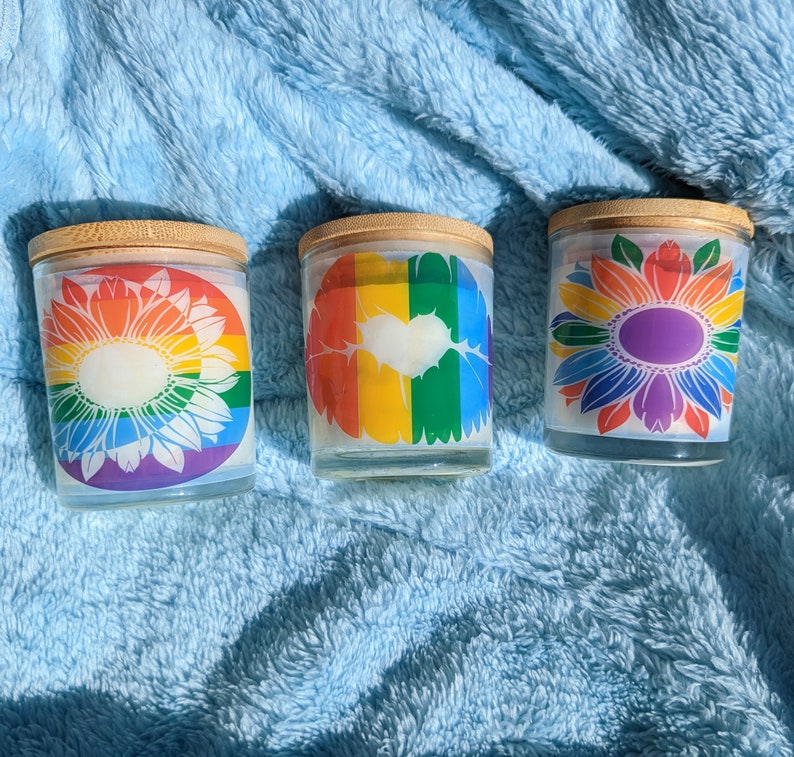 May include: Three glass candle jars with wooden lids. The jars have different designs: a rainbow with a heart, a white sunflower, and a colorful sunflower.