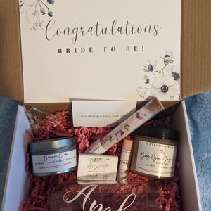 以下が含まれることがあります： 白いギフトボックスに花柄のデザインと「Congratulations Bride to Be!」の文字が書かれています。 ボックスは開いており、ピンクのティッシュペーパーで満たされています。 ボックスには、キャンドル、バスボム、石鹸、リップバーム、名前が「Amb」と書かれたグラス、そして小さなバスソルトのボトルなど、さまざまなギフトが入っています。