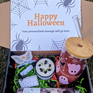 May include: A Halloween gift box filled with a variety of items, including a glass jar with a ghost design, a candle with black and green stones, a small bottle of peppermint essential oil, a salt soak, and a sticker of a cat with a third eye. The box is decorated with a spiderweb pattern and the words "Happy Halloween".