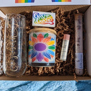 May include: A gift box filled with a rainbow candle, a glass with the word "remarkable" etched on it, a lip balm, and a tube of bath salts. The box also includes a small card with the words "Love is Love" written on it.