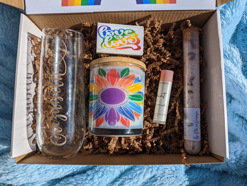 May include: A gift box filled with a rainbow candle, a glass with the word "remarkable" etched on it, a lip balm, and a tube of bath salts. The box also includes a small card with the words "Love is Love" written on it.