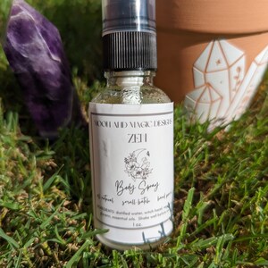 May include: A clear glass bottle with a black spray top and a white label. The label says "MOOH AHID MAGIC DESIGN ZEN Body Spray all natural small batch hand poured INGREDIENTS: distilled water, witch hazel, vegan glycerin, essential oils. Shake well before use 1 oz."