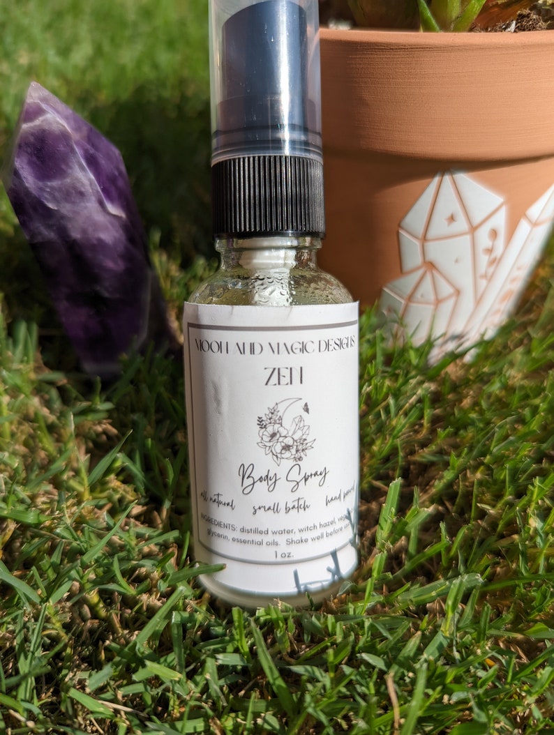 May include: A clear glass bottle with a black spray top and a white label. The label says "MOOH AHID MAGIC DESIGN ZEN Body Spray all natural small batch hand poured INGREDIENTS: distilled water, witch hazel, vegan glycerin, essential oils. Shake well before use 1 oz."