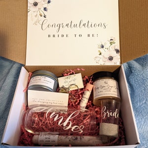 以下が含まれることがあります： 蓋に「Congratulations Bride to Be!」という文字と花柄が描かれた白いギフトボックス。ボックスが開いており、キャンドル、リング、リップバーム、シュガースクラブ、シャンパンフルート、そして「Bride」と書かれたグラスなど、さまざまなギフトが入っています。