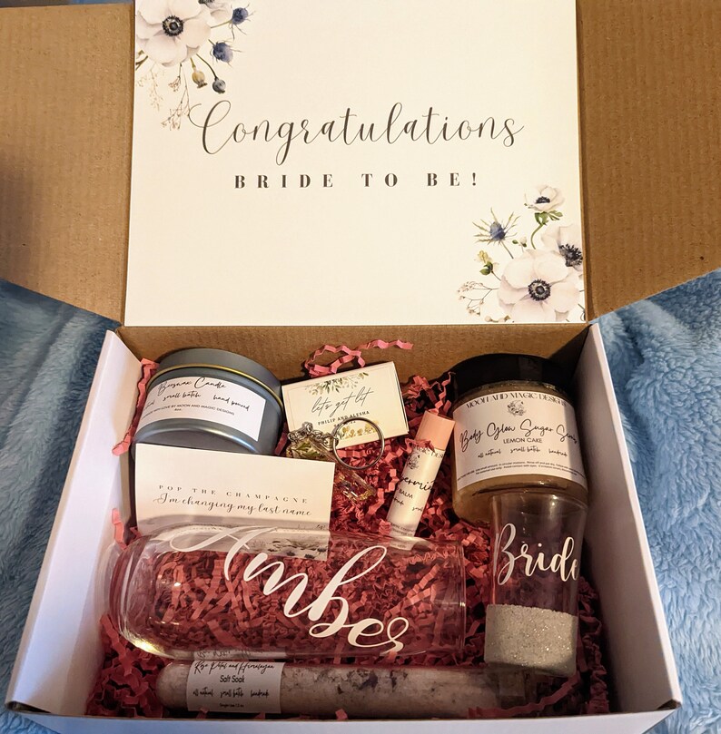 以下が含まれることがあります： 蓋に「Congratulations Bride to Be!」という文字と花柄が描かれた白いギフトボックス。ボックスが開いており、キャンドル、リング、リップバーム、シュガースクラブ、シャンパンフルート、そして「Bride」と書かれたグラスなど、さまざまなギフトが入っています。