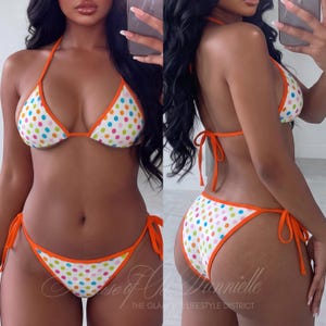 Könnte beinhalten: Ein zweiteiliger Bikini mit weißem Stoffgrund und einem bunten Punktemuster in Blau, Pink und Grün. Das Bikini-Oberteil und das Unterteil sind mit leuchtend orangefarbenem Stoff eingefasst und haben verstellbare seitliche Bänder.