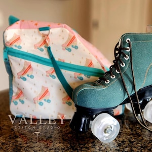 May include: A teal roller skate with brown laces and white wheels sits next to a matching bag. The bag is white with a pattern of pink and orange roller skates. The bag has a teal zipper and strap. The text "VALLEY STITCH STUDIO" is at the bottom.