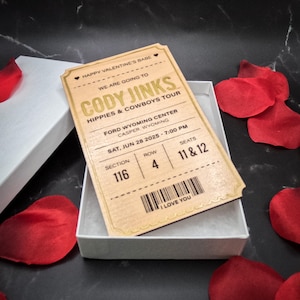 May include: A wooden ticket stub with gold lettering for a Cody Jinks concert. The ticket stub says "Happy Valentine's Babe, We are going to Cody Jinks, Hippies & Cowboys Tour, Ford Wyoming Center, Casper, Wyoming, Sat, Jun 28 2025 - 7:00 PM, Section 116, Row 4, Seats 11 & 12, I Love You".
