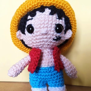 Peut inclure: Une poupée en crochet portant un chapeau jaune, un gilet rouge et un pantalon bleu. La poupée a les cheveux noirs et de grands yeux bruns.