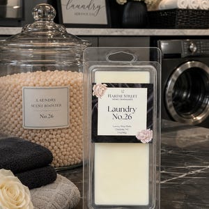 May include: A rectangular plastic clamshell package containing off-white wax melts. The label reads 12 Hardie Street Home Fragrance Laundry No.26 Luxury Wax Melts Charlotte, NC 1.7oz/48g, decorated with pink floral illustrations on a black background.