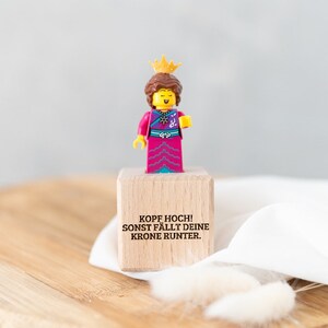 Könnte beinhalten: Ein kleiner Holzblock mit dem Text "Kopf hoch! Sonst fällt deine Krone runter." Eine Lego-Figur einer Königin in einem rosa-blauen Kleid und einer goldenen Krone steht auf dem Block.