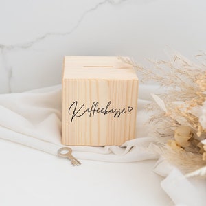 Könnte beinhalten: Eine Holzkasse mit einem Schlitz oben und dem Text "Kaffekasse" mit einem Herzsymbol. Die Kasse steht auf einer weißen Oberfläche mit einem Strauß getrockneter Blumen.