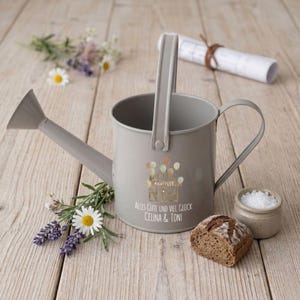 Puede incluir: Una regadera de metal gris con asa y boquilla, decorada con un gráfico de globos y texto. La regadera está rodeada de flores, un pequeño cuenco de sal y un trozo de pan sobre una superficie de madera.