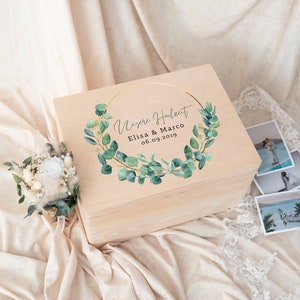 Pu&ograve; includere: Una scatola di legno color chiaro con un design circolare dorato e accenti di foglie verdi. Il testo "Unsere Hochzeit" &egrave; sopra i nomi "Elisa & Marco" e la data "06.09.2019". Un piccolo bouquet e foto sono accanto alla scatola.
