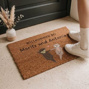 May include: A rectangular doormat made of natural brown fibers. The mat features the text "WILLKOMMEN BEI Moritz und Antonia" in black, along with illustrations of two otters holding hands. A small vase with dried flowers is visible.