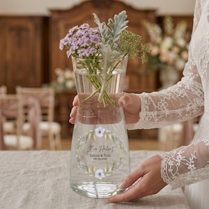 Könnte beinhalten: Eine klare Glasvase mit Blumengirlanden-Design und Text, die einen Strauß aus lila und grünen Blumen enthält. Die Vase ist mit Wasser gefüllt. Der Text lautet "Zur Hochzeit Celina & Toni 06.09.2037".