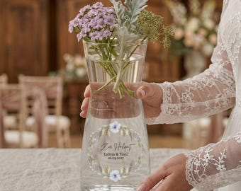 May include: A clear glass vase filled with water and flowers, featuring a floral wreath design with text. The vase holds a bouquet of purple and green flowers. The text reads "Zur Hochzeit Celina & Toni 06.09.2037".
