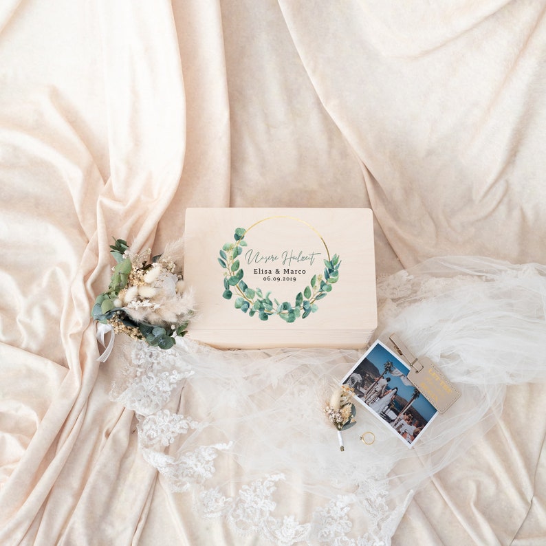 Pu&ograve; includere: Una scatola di legno color chiaro con un design a ghirlanda color oro e testo. La scatola &egrave; decorata con una composizione floreale, una fotografia, un anello e un velo. Il testo sulla scatola recita "Unsere Hochzeit Elisa & Marco 06.09.2019".