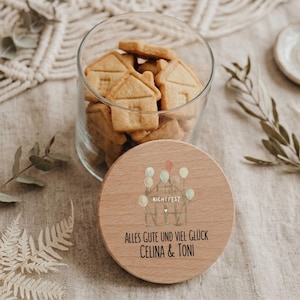 Op de afbeelding: Een glazen pot gevuld met huisvormige koekjes, met een houten deksel met een ballon- en huisafbeelding. Op het deksel staat de tekst "Alles Gute und Viel Glück Celina & Toni."