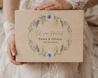 Puede incluir: Una caja de madera de color claro con un diseño de corona floral. La corona presenta flores azules y hojas verdes. El texto "Unsere Hochzeit" está encima de los nombres "Paula & Johann" y la fecha "20.03.2023".