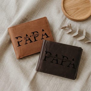 Könnte beinhalten: Zwei Ledergeldbörsen, eine hellbraune und eine dunkelbraune, werden gezeigt. Das Wort "PAPA" ist in großen, fetten Buchstaben auf jeder Geldbörse aufgedruckt. Auf der hellbraunen Geldbörse stehen die Namen "Julia Matteo" unter dem Wort "PAPA". Auf der dunkelbraunen Geldbörse stehen die Namen "Clara Timan Matilde".