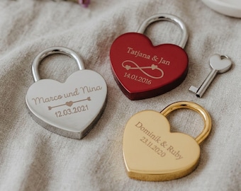 Può includere: Tre lucchetti a forma di cuore in argento, rosso e oro, ciascuno inciso con nomi e date. Una chiave d'argento è presente. Il lucchetto d'argento recita "Marco und Nina 12.03.2021". Il lucchetto rosso recita "Tatjana & Jan 14.01.2016". Il lucchetto d'oro recita "Dominik & Ruby 23.11.2020".