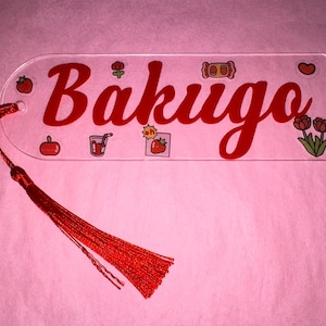 May include: A clear acrylic bookmark with the name "Bakugo" in red script. The bookmark is decorated with small, colorful illustrations of food and flowers. A red tassel hangs from the top.