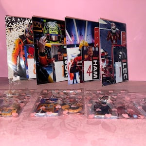 Puede incluir: Una exhibici&oacute;n de merchandising con tem&aacute;tica de carreras de F&oacute;rmula 1. La imagen presenta varias fotos enmarcadas de pilotos de carreras y paquetes de pl&aacute;stico transparente que contienen llaveros con dise&ntilde;os de coches de carreras. El fondo es rosa liso.