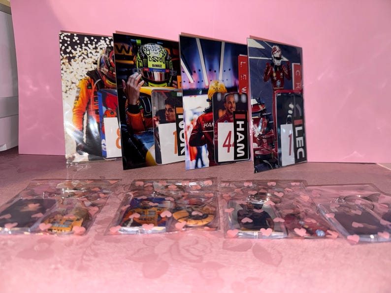 Puede incluir: Una exhibici&oacute;n de merchandising con tem&aacute;tica de carreras de F&oacute;rmula 1. La imagen presenta varias fotos enmarcadas de pilotos de carreras y paquetes de pl&aacute;stico transparente que contienen llaveros con dise&ntilde;os de coches de carreras. El fondo es rosa liso.