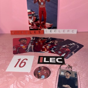 Puede incluir: Una colecci&oacute;n de recuerdos de Charles Leclerc, que incluye una tarjeta fotogr&aacute;fica, pegatinas, un bot&oacute;n y un llavero. Los art&iacute;culos presentan im&aacute;genes de Leclerc con su traje de carreras y el n&uacute;mero 16. El fondo es un estampado floral rosa.