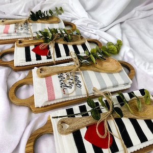May include: Four wooden cutting boards with handles, each wrapped with a tea towel and a wooden spoon. The tea towels are striped in black and white, gray and white, or red and white. The tea towels have embroidered sayings like "Love the Kitchen" and "Home is where the food is".