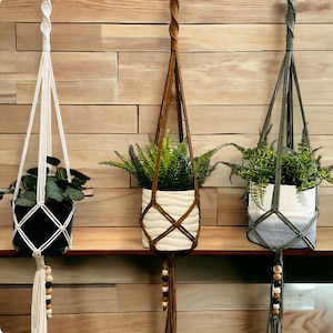 May include: Three macrame plant hangers with wooden beads hanging from a wooden shelf. The hangers are made with white, brown, and green cord. Each hanger holds a potted plant.