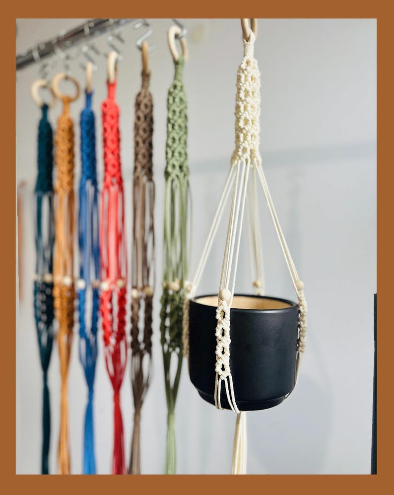 May include: A collection of macrame plant hangers in various colors, including teal, blue, red, brown, and green, are displayed. A black ceramic pot is suspended in a cream-colored macrame hanger. The hangers are designed to hold potted plants.