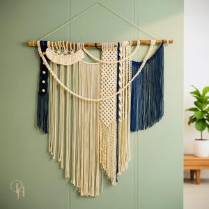 May include: Macrame wall hanging featuring a natural wooden dowel and a variety of off-white and navy blue knotted cotton cords. The design includes fringe, geometric patterns, and decorative wooden beads. This handcrafted piece adds a bohemian touch to any space.