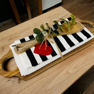 May include: A wooden serving tray with a black and white striped kitchen towel, a wooden spoon, and a red heart. The towel is tied with twine and a sprig of greenery.