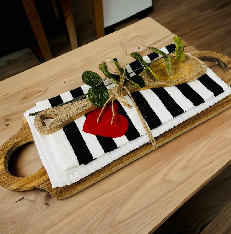 May include: A wooden serving tray with a black and white striped kitchen towel, a wooden spoon, and a red heart. The towel is tied with twine and a sprig of greenery.