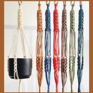 May include: A collection of macrame plant hangers in various colors, including cream, orange, blue, red, brown, green, and teal. Each hanger features a wooden ring and beads, designed to suspend a plant pot. A black ceramic pot is shown in one hanger.