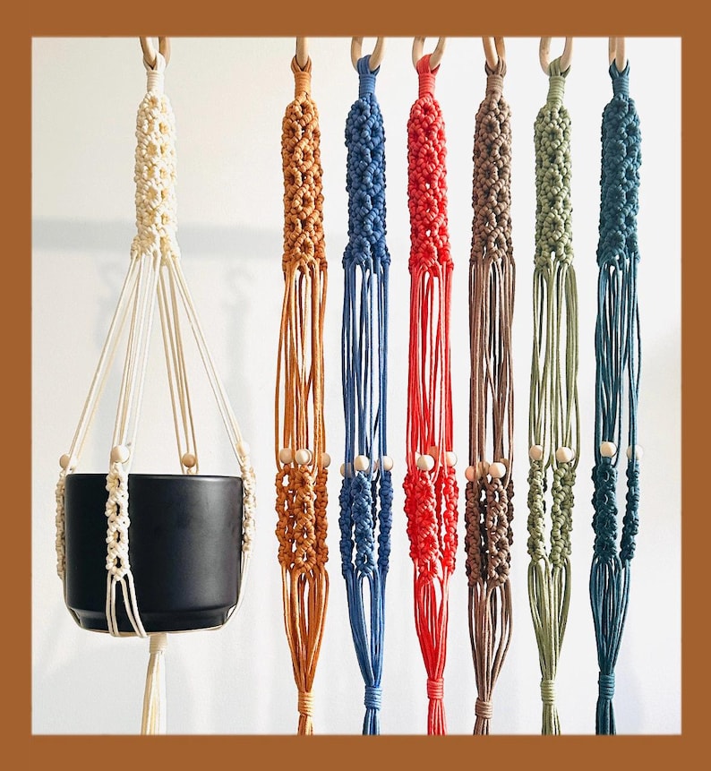 May include: A collection of macrame plant hangers in various colors, including cream, orange, blue, red, brown, green, and teal. Each hanger features a wooden ring and beads, designed to suspend a plant pot. A black ceramic pot is shown in one hanger.