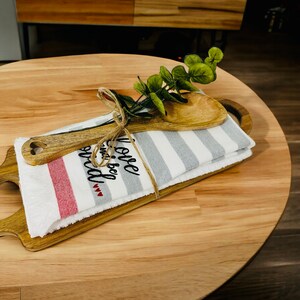 May include: A wooden serving tray with a white and gray striped kitchen towel and a wooden spoon. The towel has the text "Love and be wild" embroidered on it. A sprig of green eucalyptus is tied to the towel with twine.