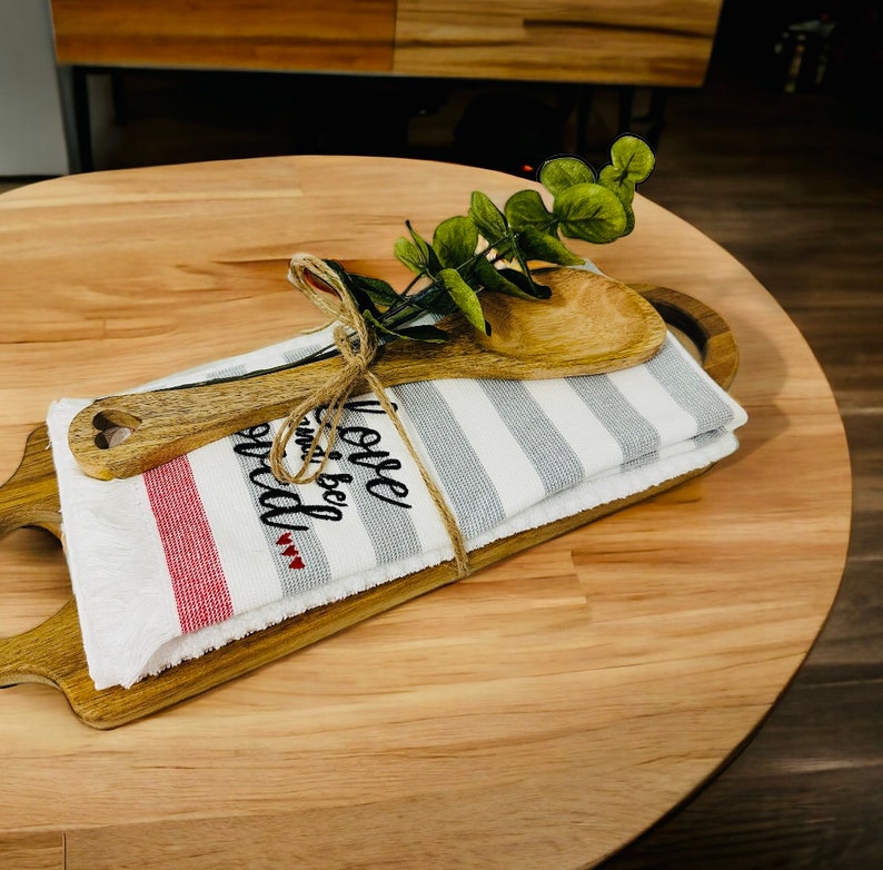 May include: A wooden serving tray with a white and gray striped kitchen towel and a wooden spoon. The towel has the text "Love and be wild" embroidered on it. A sprig of green eucalyptus is tied to the towel with twine.