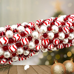 May include: A close-up of a red and white candy cane bead garland. The garland is made of white beads with red stripes and red beads. The garland is on a wooden surface with a blurry background of Christmas lights.