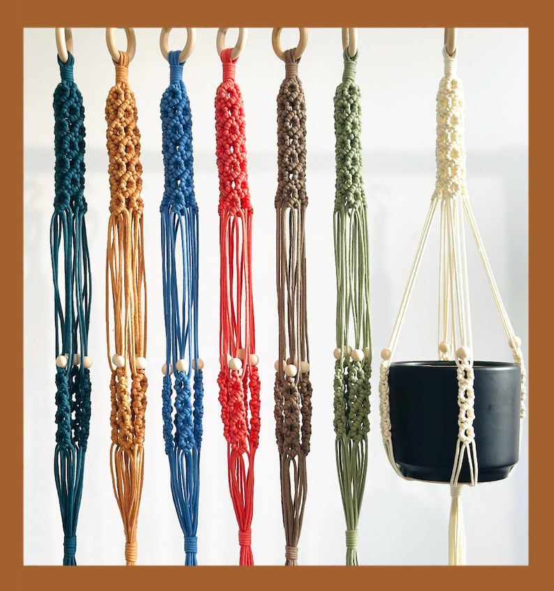 May include: A collection of macrame plant hangers in various colors, including teal, tan, blue, red, brown, olive green, and cream. Each hanger features a wooden ring at the top and wooden beads. One hanger holds a black ceramic pot.