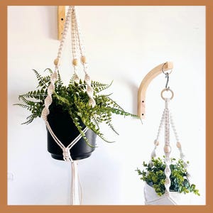 May include: Two macrame plant hangers with wooden beads, one hanging from a wooden wall mount. The plant hangers are white and feature a black pot with green plants.