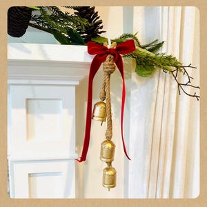 May include: A decorative Christmas bell ornament. Three gold-colored bells hang from a natural rope, adorned with a red velvet ribbon. The bells are suspended from a knot, with a red ribbon bow at the top. The ornament is part of a holiday decoration.
