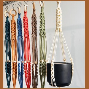 May include: A collection of macrame plant hangers in various colors, including teal, orange, blue, red, brown, olive green, and cream. Each hanger features a wooden ring at the top and wooden beads. A black ceramic pot is suspended in one of the cream-colored hangers.