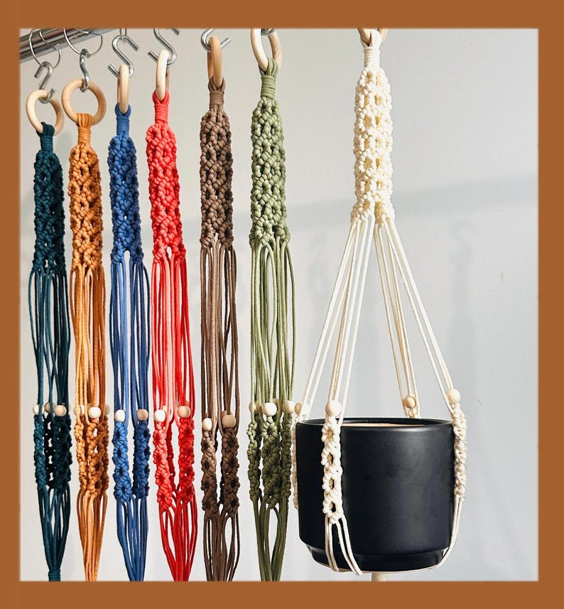 May include: A collection of macrame plant hangers in various colors, including teal, orange, blue, red, brown, olive green, and cream. Each hanger features a wooden ring at the top and wooden beads. A black ceramic pot is suspended in one of the cream-colored hangers.