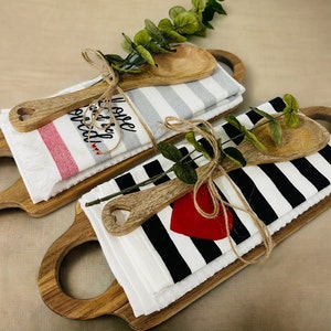 May include: Two wooden cutting boards with handles, each with a white kitchen towel, a wooden spoon, and twine. One towel has red and white stripes and says "I love you more than bread". The other towel has black and white stripes and a red heart.